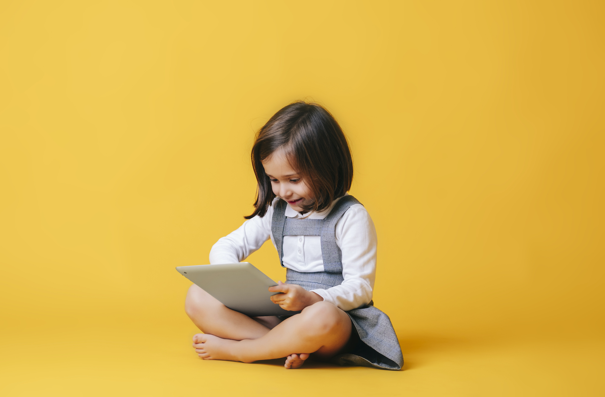 A caucasian girl in a gray dress and white shirt uses and plays with a tablet on a yellow background